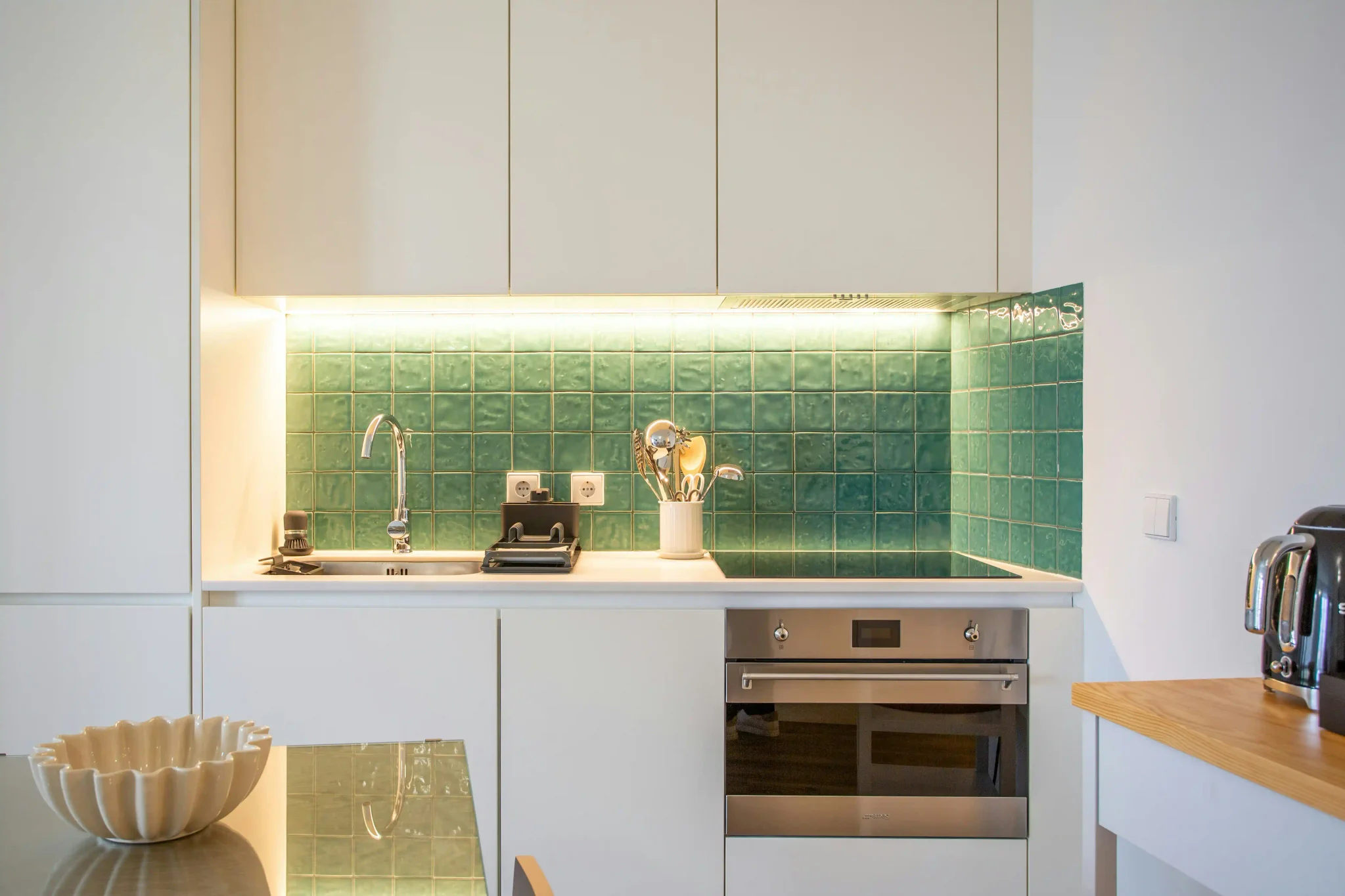 White kitchen with glossy green zellige square tile backsplash lit by under-cabinet lighting