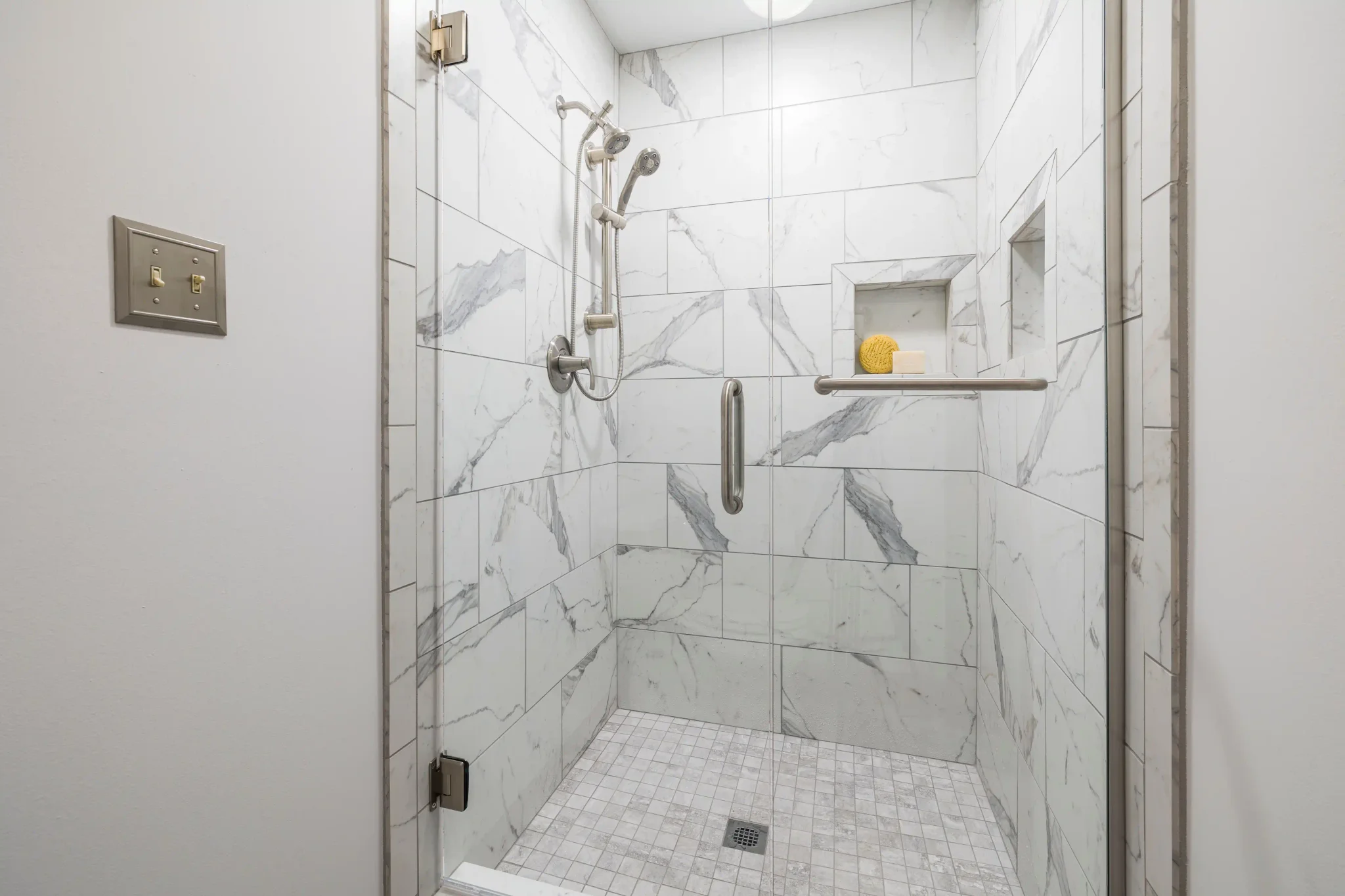 Walk-in shower with white marble-look tile, glass door, grab bar, and built-in niche